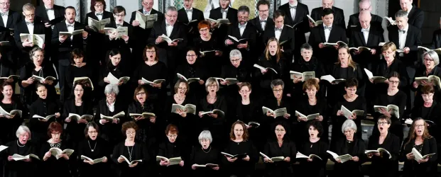 Ein großer Chor mit vielen Sängern in schwarzen Kleidungen steht zusammen auf einer Bühne. Alle halten Notenblätter und singen gemeinsam.