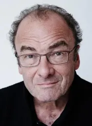 Ein Mann mit Brille und leichtem Lächeln schaut in die Kamera. Er hat graues Haar und trägt ein schwarzes Oberteil.