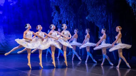 Eine Gruppe von Ballerinen in weißen Tutus tanzt auf einer Bühne. Der Hintergrund ist blau beleuchtet, was eine elegante Atmosphäre schafft.