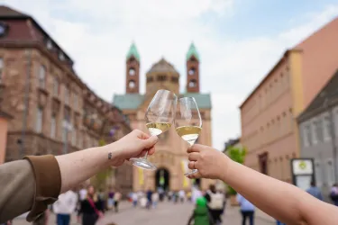 Zwei Personen stoßen mit Gläsern Sekt an, während im Hintergrund eine historische Kirche zu sehen ist. Der Platz ist belebt und es herrscht eine festliche Stimmung.