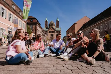 Eine Gruppe junger Menschen sitzt auf einem Pflastersteinplatz und genießt die Sonne. Im Hintergrund ist eine große Kirche und historische Gebäude zu sehen.