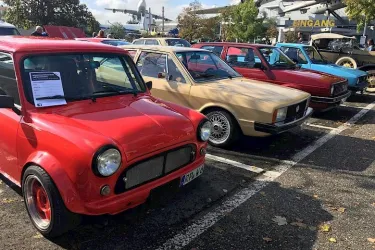 Eine Reihe von klassischen Autos in verschiedenen Farben steht auf einem Parkplatz. Im Vordergrund ist ein rotes Auto zu sehen.
