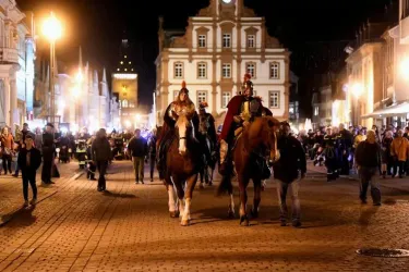 Zwei Reiter in historischen Kostümen reiten auf Pferden durch eine belebte Straße bei Nacht. Im Hintergrund sind Menschen und beleuchtete Gebäude zu sehen.