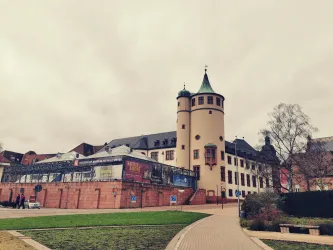 Ein historisches Gebäude mit einem runden Turm und einer spitzen Dachspitze. Der Himmel ist bewölkt und die Umgebung ist grün.