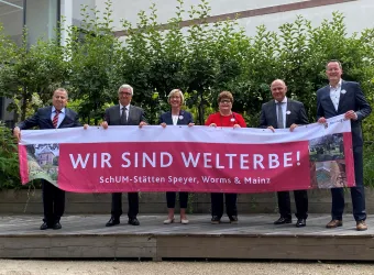 Sechs Personen stehen auf einer Bühne und halten ein großes Banner mit der Aufschrift „WIR SIND WELTERBE!“. Im Hintergrund sind grüne Pflanzen zu sehen.