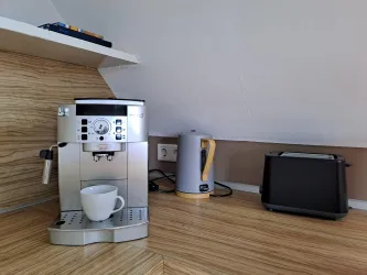 Eine moderne Küchenecke mit einer Espressomaschine, einem Wasserkocher und einem Toaster. Der Boden ist aus hellem Holz, und ein weißer Becher steht bereit.