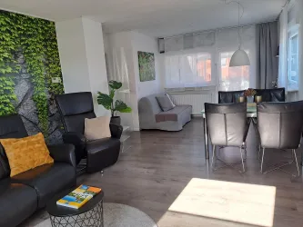Ein modernes Wohnzimmer mit bequemen schwarzen Sesseln und einem grauen Sofa. Der Raum hat große Fenster, die viel Tageslicht hereinlassen, sowie eine dekorative grüne Wand.