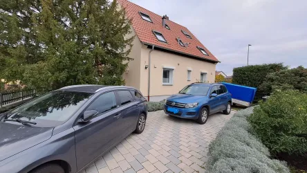 Ein Parkplatz mit zwei Autos, einem grauen Fahrzeug und einem blauen Anhänger. Im Hintergrund ist ein Haus mit rotem Dach und Bäumen zu sehen.