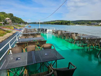 Ein Schiff mit vielen Tischen und Stühlen auf dem Deck. Im Hintergrund fließt ein ruhiger Fluss, umgeben von Bäumen und Hügeln.