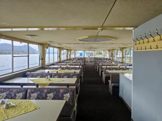 Ein gemütlicher Speiseraum mit Tischen und Stühlen. Große Fenster bieten einen Blick auf das Wasser.