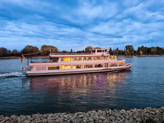 Ein Boot fährt gemütlich auf einem Fluss unter einem bewölkten Himmel. Die Lichter des Bootes spiegeln sich im Wasser.