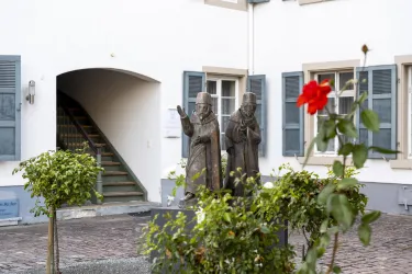 Zwei bronzene Statuen stehen in einem Innenhof mit grünen Pflanzen. Im Vordergrund blüht eine rote Rose.