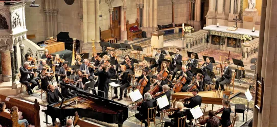 Ein Orchester spielt in einer Kirche. Die Musiker sind konzentriert und es herrscht eine festliche Atmosphäre.