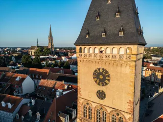 Ein historischer Stadtturm mit einer großen Uhr und einem klaren Himmel. Im Hintergrund sind weitere Dächer und Kirchtürme sichtbar.