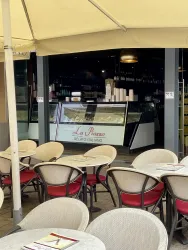 Ein einladendes Café mit Stühlen und Tischen im Freien. Im Hintergrund ist eine Eistheke mit der Aufschrift "La Piazza" zu sehen.