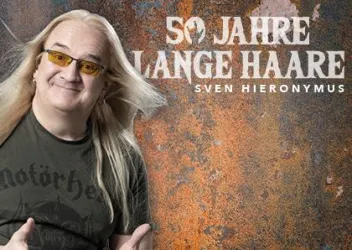 Ein lächelnder Mann mit langen, blonden Haaren trägt ein schwarzes T-Shirt. Im Hintergrund steht der Schriftzug „50 Jahre lange Haare“.