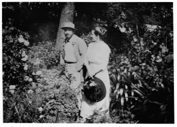 Ein Paar steht in einem Garten umgeben von Blumen. Der Mann trägt einen Hut und die Frau eine helle Kleidung mit einem Hut.
