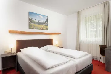 Ein helles und gemütliches Schlafzimmer mit zwei Einzelbetten. An der Wand hängt ein Bild einer Landschaft, und ein Fenster lässt viel Tageslicht herein.
