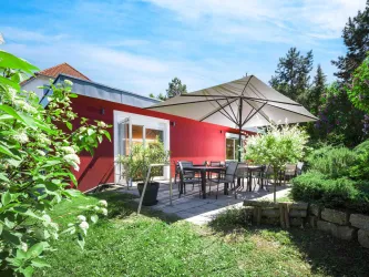 Eine moderne rote Villa mit einer Terrasse und einem großen Sonnenschirm. Der Garten ist grün und einladend, ideal zum Entspannen.