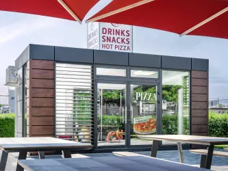 Ein moderner Snackstand mit einem großen Schild, das "Drinks, Snacks, Hot Pizza" anzeigt. Draußen stehen Tische unter roten Sonnenschirmen.