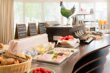 Ein Buffet mit verschiedenen Speisen wie Aufschnitt, Käse und frischem Obst. Im Hintergrund steht eine dekorative Henne und die Tische sind ordentlich gedeckt.