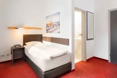Ein modernes Einzelzimmer mit rotem Teppich und einfacher Ausstattung. Es gibt ein Bett, einen Nachttisch und ein Kunstwerk an der Wand.