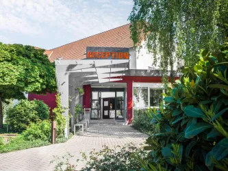 Ein Empfangsbereich mit großen Fenstern und einem modernen Eingang. Umgeben von üppigem Grün und Pflanzen.