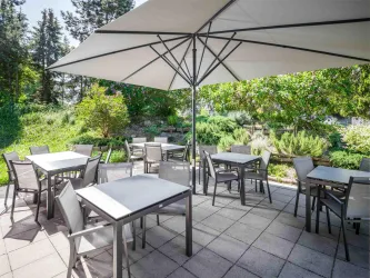 Ein gemütlicher Außenbereich mit weißen Tischen und Stühlen unter einem großen Sonnenschirm. Der grüne Garten im Hintergrund sorgt für eine angenehme Atmosphäre.