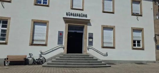 Ein helles Verwaltungsgebäude mit dem Schriftzug "Bürgerhaus" über dem Eingang. Vor dem Gebäude stehen eine Bank und ein Fahrrad.
