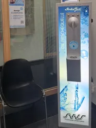 Ein Wasserspender mit einer klaren Anzeige und einem modernen Design steht in einem Raum. Daneben befindet sich ein schwarzer Stuhl.
