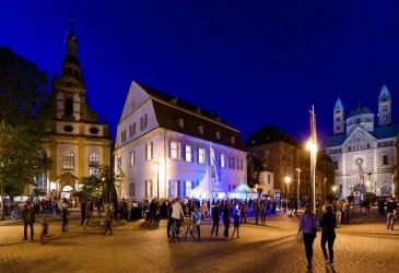 Eine lebhafte Abendveranstaltung auf einem Platz mit mehreren Gebäuden im Hintergrund. Die Szene ist mit vielen Menschen und festlicher Beleuchtung belebt.