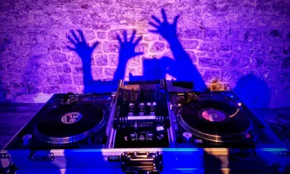 Eine DJ-Konsole mit Schallplatten und einem Mischpult. Die bunten Schatten von Händen sind an der Wand zu sehen.