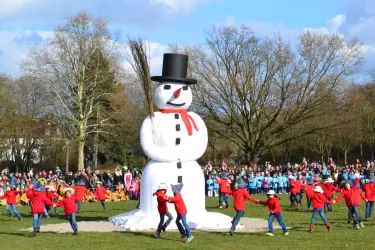Ein großer Schneemann steht in einem Park, während Kinder in roten Jacken um ihn herum tanzen. Im Hintergrund sind fröhliche Zuschauer und Bäume zu sehen.