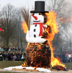 Ein großer Schneemann brennt lodernd in einem Feld. Im Hintergrund stehen Menschen und schauen zu.