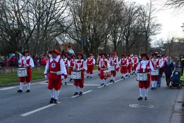 Eine Gruppe von Trommlern in roten Kostümen marschiert auf einer Straße. Zuschauer stehen an der Seite und beobachten den Umzug.