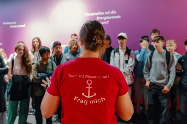 Eine Gruppe von Jugendlichen hört einer Person in einem roten T-Shirt mit der Aufschrift „Frag mich“ zu. Der Hintergrund zeigt verschiedene Fragen zur Energieversorgung.