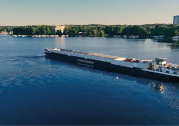 Ein großes Frachtschiff fährt auf einem ruhigen Gewässer. Im Hintergrund sind Bäume und Gebäude zu sehen.