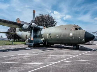 Ein militärisches Transportflugzeug steht auf einem Parkplatz. Der Himmel ist blau mit einigen Wolken.