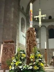 Ein dekorierter Altar mit einer großen Christuskerze und einem Kreuz im Hintergrund. Blumenarrangements ergänzen die festliche Stimmung in der Kirche.
