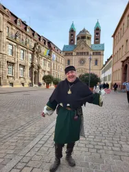 Ein lächelnder Mann in historischer Kleidung steht auf einer gepflasterten Straße. Im Hintergrund ist ein beeindruckendes historisches Gebäude zu sehen.
