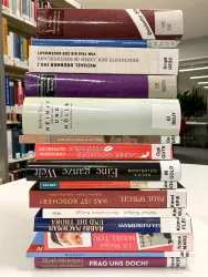 Ein Stapel von bunten Büchern steht auf einem Tisch in einer Bibliothek. Die Buchrücken sind gut sichtbar und zeigen verschiedene Titel und Farben.