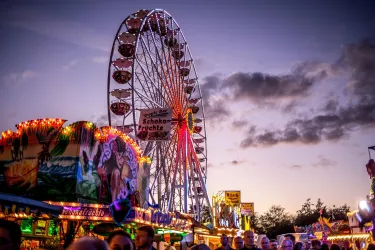 Ein Riesenrad leuchtet am Abendhimmel, umgeben von bunten Fahrgeschäften und festlicher Stimmung. Viele Menschen genießen den Spaß auf dem Jahrmarkt.