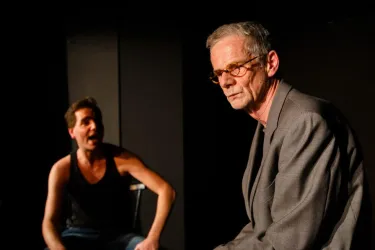 Zwei Männer auf einer Theaterbühne. Der Mann links wirkt lebhaft und emotional, während der Mann rechts ernst und nachdenklich aussieht.