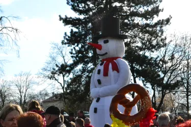 Eine große Schneemann-Figur mit einem Hut und einer Karottennase steht inmitten einer Menschenmenge. Im Vordergrund ist eine Brezel zu sehen.