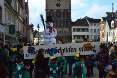 Eine fröhliche Parade mit Kindern in bunten Kostümen und einem großen Schneemann. Im Hintergrund sieht man das Stadtgebäude und eine festliche Atmosphäre.