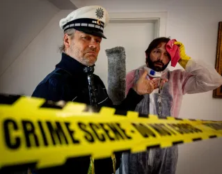 Ein Polizist und ein Mann in Schutzkleidung stehen an einem Tatort. Gelbe Absperrbänder mit der Aufschrift "CRIME SCENE - DO NOT CROSS" sind im Hintergrund sichtbar.