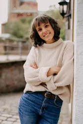 Eine lächelnde Frau steht an einer Wand und trägt einen beigen Pullover und Jeans. Im Hintergrund sind Bäume und eine alte Gebäudestruktur sichtbar.
