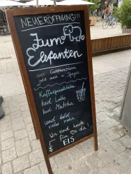 Ein schwarzes Schild mit der Aufschrift „Elefanten“ und Informationen zu Speisen und Getränken. Es gibt einen Hinweis auf die Neueröffnung und verschiedene Angebote.