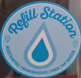 Ein runder Aufkleber mit dem Text „Refill Station“ und einem Wassertropfen darauf. Darunter steht „Kostenfrei Trinkwasser / Free Tap Water“.
