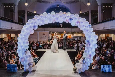 Eine Modenschau mit einem Brautpaar auf dem Laufsteg unter einem Ballonbogen. Die Menge im Hintergrund schaut begeistert zu.
